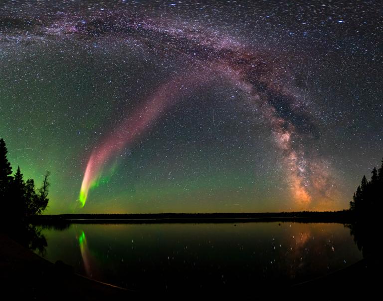 A vivid arc of pinkish-red and green aurora curves across a starry night sky alongside the Milky Way's core, above a dark tree-lined lake that mirrors the lights on its calm surface. Partially obscured.