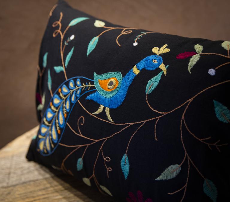 Close-up photo of a rectangular black throw pillow resting on a wooden bench, featuring detailed embroidery of a stylized peacock with a curved blue neck, teal head crest, and an orange-and-teal eye-patterned feather. The design also includes brown vine-like stitching, teal and sage leaves, and small maroon and white floral accents scattered across the black fabric. Partially obscured.