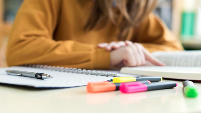Close-up of a spiral notebook, a pen, and colorful highlighters on a desk, with a person in a mustard sweater sitting blurred in the background. Partially obscured.