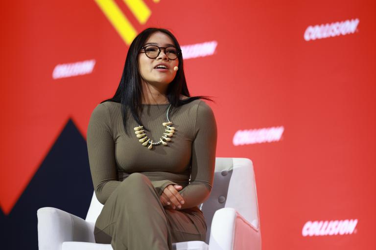 A woman wearing glasses and a statement necklace speaks into a headset microphone, with a red and yellow graphic backdrop behind her. Partially obscured.