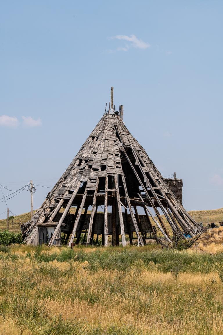 A large, weathered wooden structure shaped like a tepee stands in a dry grassy field under a clear blue sky. The structure's wooden frame is largely exposed, with many wood shingles missing or falling off, revealing the ribbed skeleton beneath. Partially obscured.