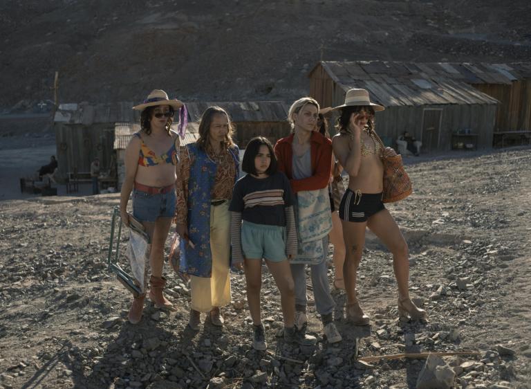 Five members of a queer family stand together on a rocky hillside in front of weathered wooden shanties in a desert mining town, with a young girl at the centre of the group looking in the same direction just off-camera. Partially obscured.