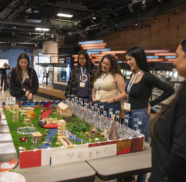 A group of students stand around a diorama displayed on a table in a museum gallery. Partially obscured.