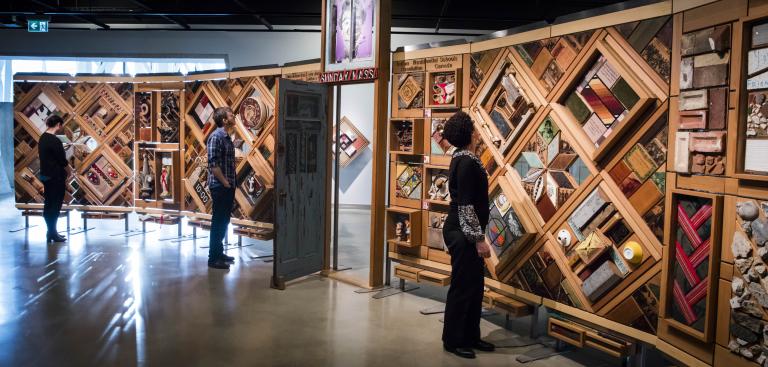 Three people in a gallery examining the Witness Blanket, a large free-standing blanket made of cedar panels holding objects related to the residential school experience. Partially obscured.