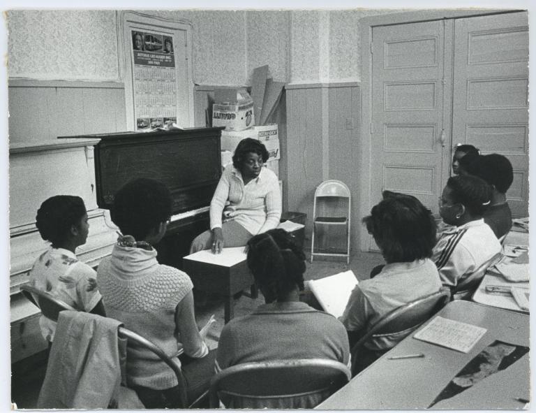 A black and white photo of a Black woman seated on a piano bench, twisted three-quarters towards a group of Black students. The students are seated in a semicircle in metal folding chairs and are holding notebooks. A table with two pens, a notebook with the top page filled with writing and a ripped piece of a poster is behind the students. Partially obscured.