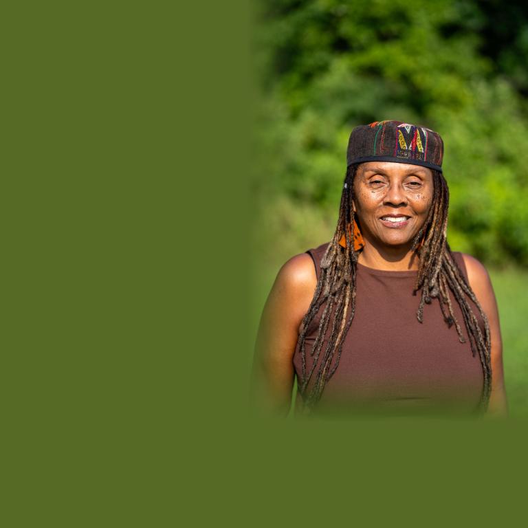 Outdoor portrait of a smiling woman with long gray-streaked locs, wearing a patterned kufi-style cap, orange patterned earrings, and a brown sleeveless top, with a soft green foliage background. Partially obscured.