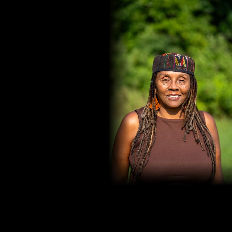 Outdoor portrait of a smiling woman with long gray-streaked locs, wearing a patterned kufi-style cap, orange patterned earrings, and a brown sleeveless top, with a soft green foliage background. Partially obscured.