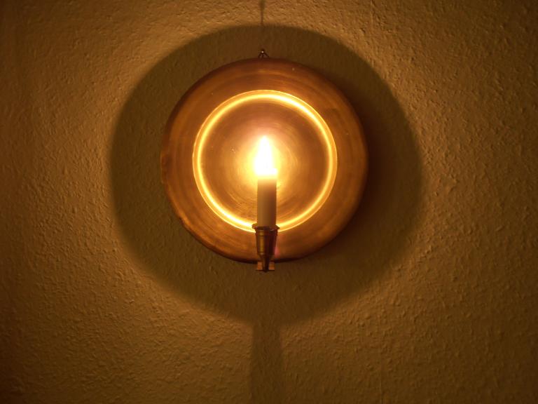 Lit candle on a round brass wall sconce, its reflector glowing gold and casting a circular shadow on a textured wall. Partially obscured.