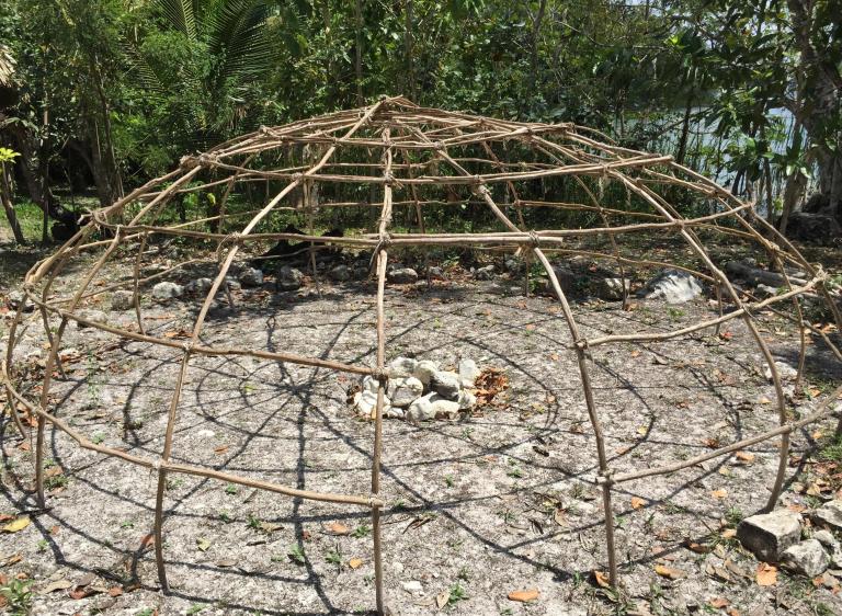 A dome frame of lashed wooden poles over a central pile of stones, surrounded by trees with water beyond. Partially obscured.