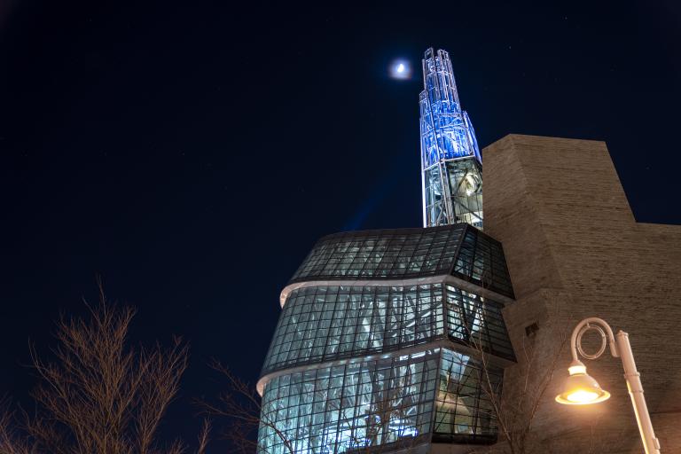 The Canadian Museum for Human Rights at night, its blue-lit Tower of Hope rising beside a crescent moon in a starry sky. Partially obscured.