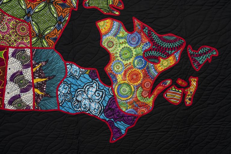 Quilt detail of a map of eastern Canada, each region cut from a bright patterned fabric and outlined in red on black. Partially obscured.