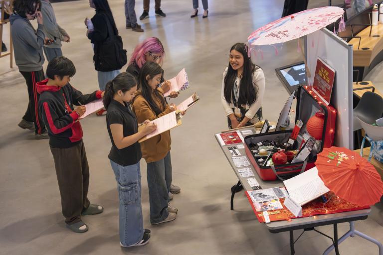 A student stands beside a table on which her project is displayed. Four students stand in front of her writing on clipboards. Partially obscured.