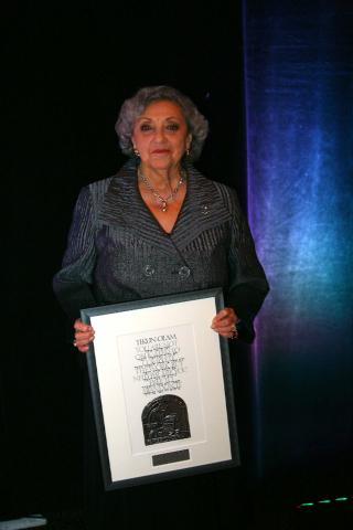 A woman is smiling at the camera and holding up a framed certificate that says “Tikun Olam. You are not obligated to complete the work but neither are you free to abandon it.