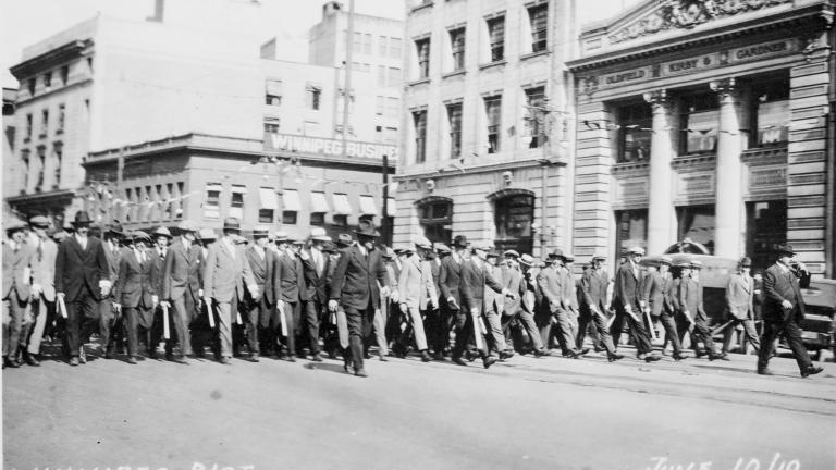 A large group of men in suits and hats brandishing clubs marching down the street.
